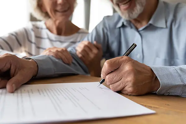 smiling older, mature man and woman sit at desk while man puts his signature on a document